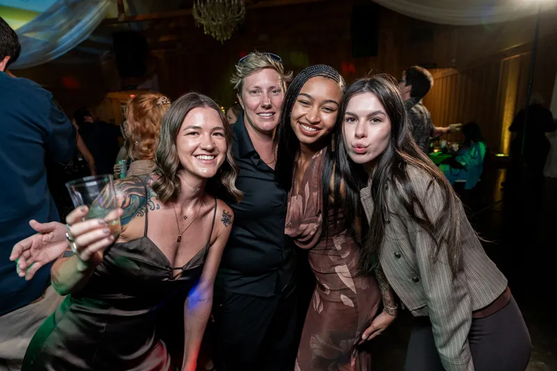 Four women smile together at the reception, wearing pink, black, and brown tones.