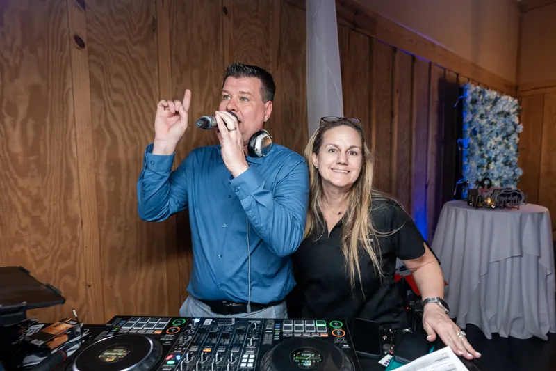 DJ in blue shirt and woman in black at mixing equipment during reception.