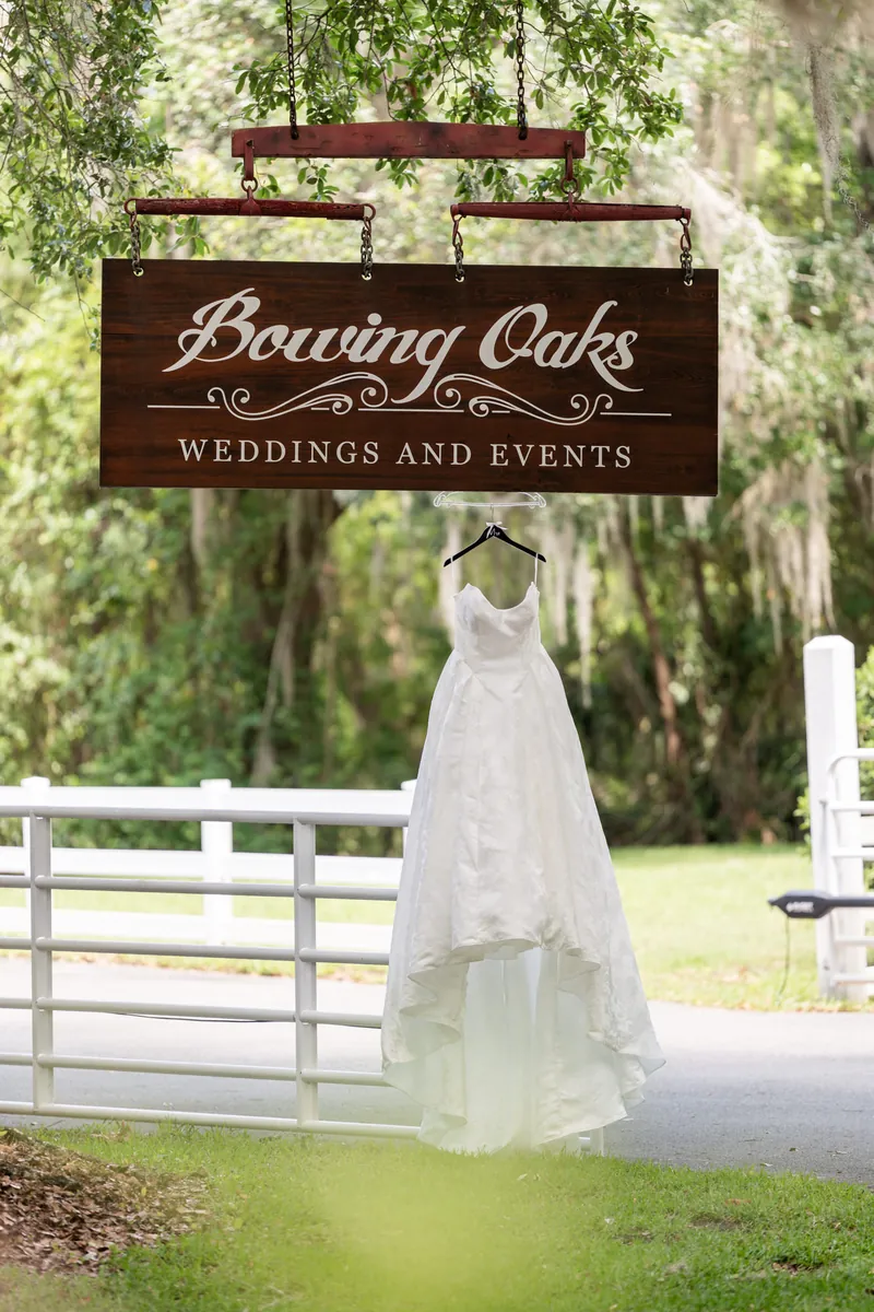 Giselle's lace wedding dress drapes beneath the Bowing Oaks wedding venue sign under sprawling oak trees.