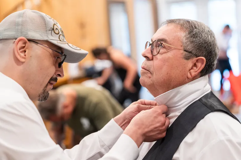 A man adjusts another man's tie indoors during wedding preparations.