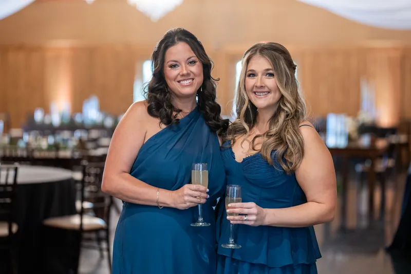 Two women in blue sleeveless dresses hold champagne glasses at an indoor reception venue.