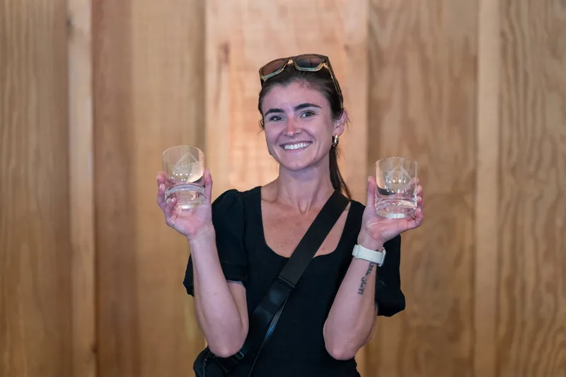 Woman in black V-neck shirt holding two glasses of pink drink, smiling at camera indoors.
