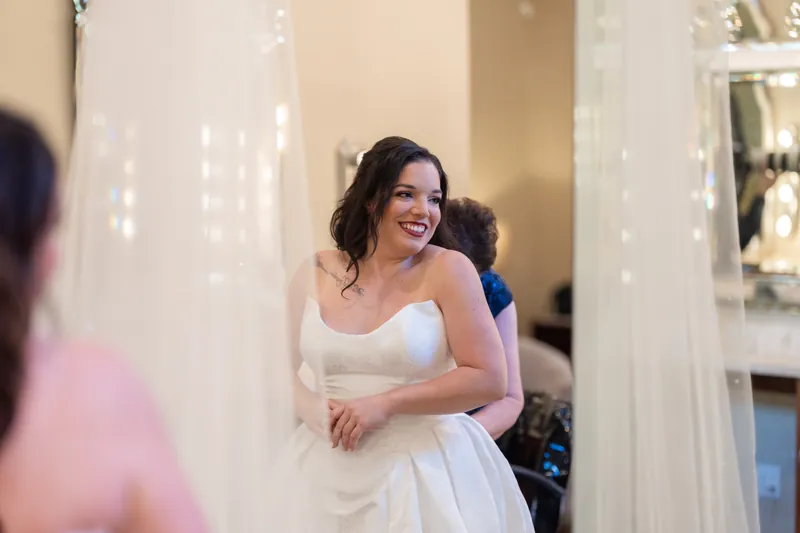 Giselle in white strapless wedding dress smiles while adjusting her veil in a bridal suite.
