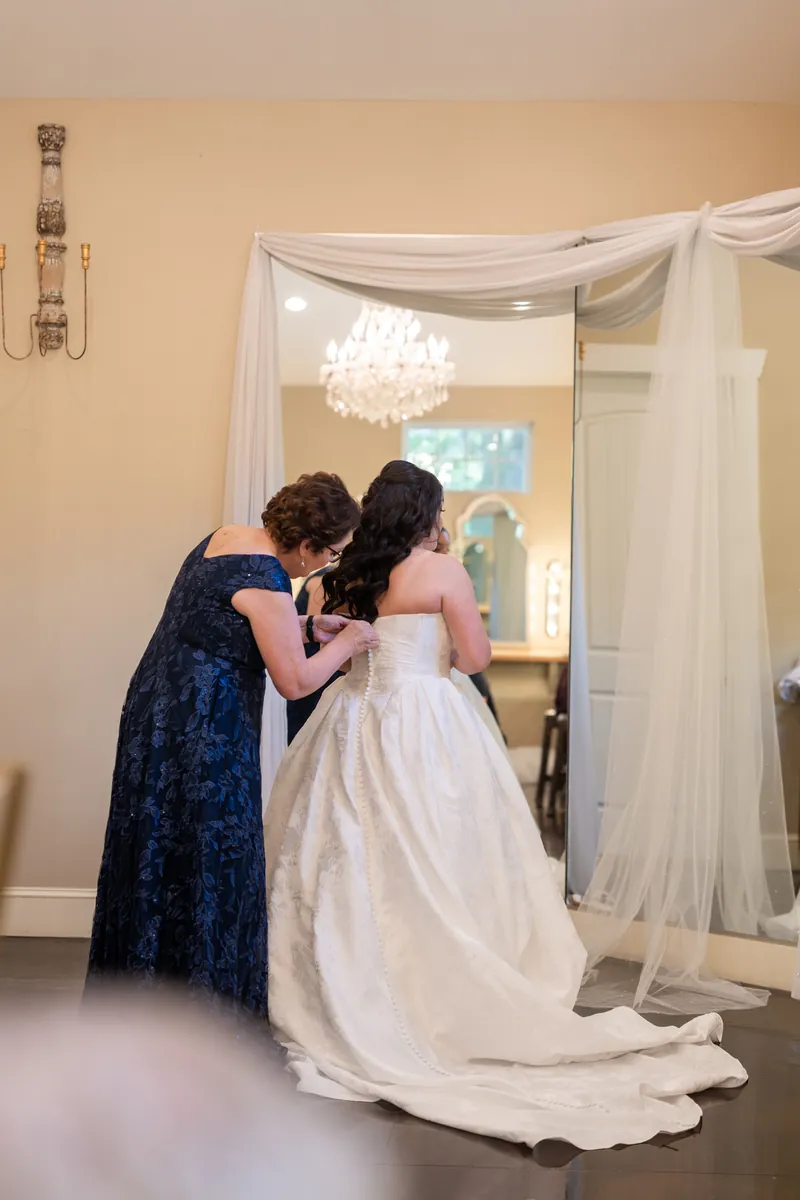 Guest in dark blue dress embraces Giselle from behind as she adjusts her white wedding gown.