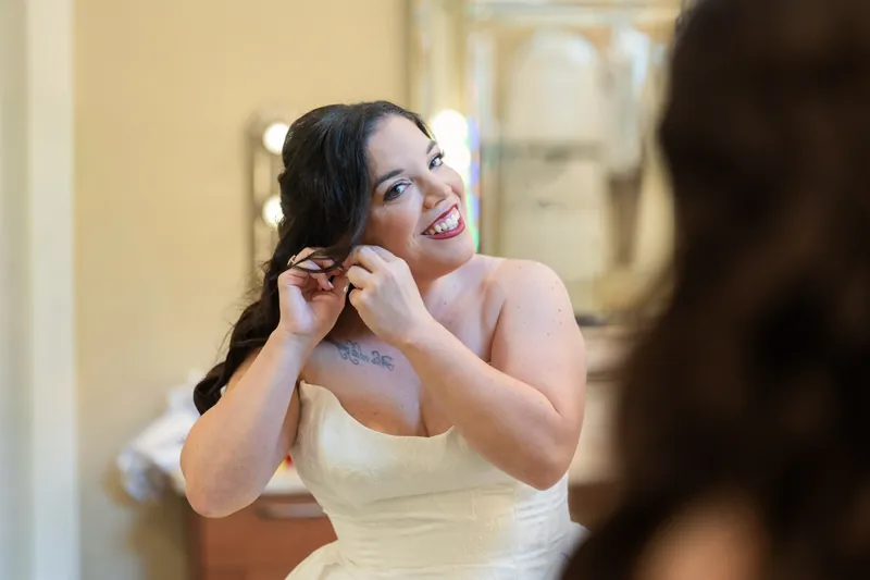 Giselle fastens an earring while wearing her white wedding dress, preparing for ceremony.
