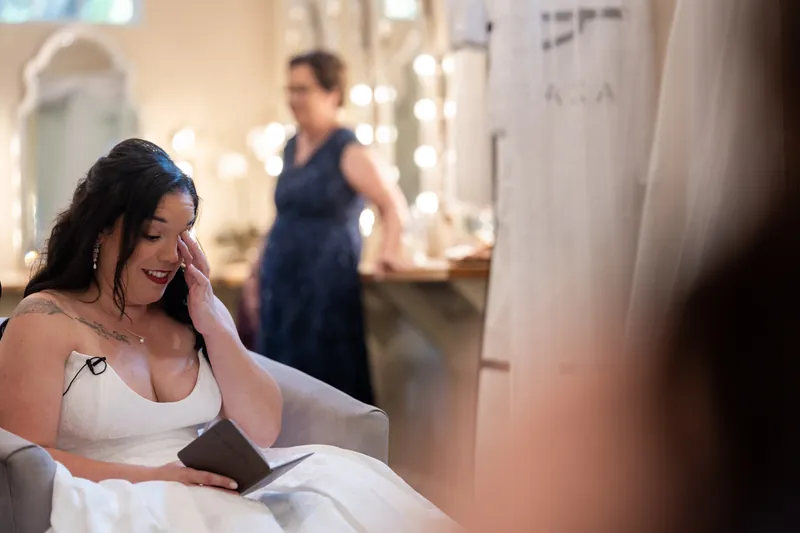 Giselle sits in a white wedding dress reading from a notebook in a bridal suite with vanity mirrors.