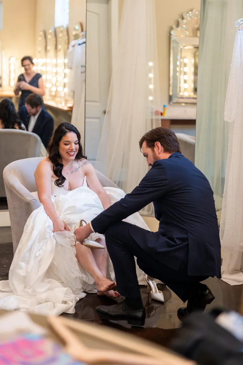 Duncan kneels beside Giselle's chair to help fasten her wedding shoe in the bridal dressing room.