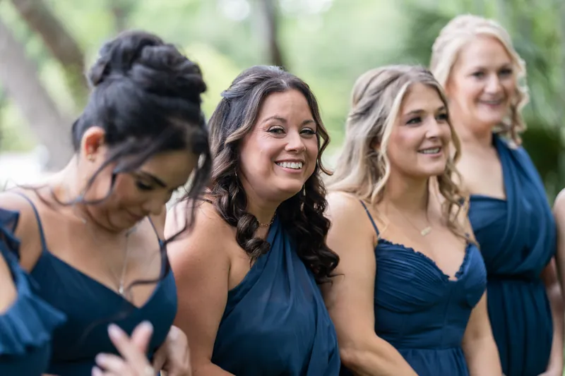Four bridesmaids in navy blue wrap dresses smile together outdoors at the wedding venue.