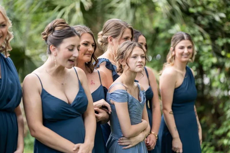 Bridesmaids in navy blue dresses stand together among green foliage during outdoor wedding celebrations.