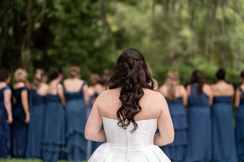 Giselle in strapless white wedding dress faces bridesmaids in blue gowns under tree-lined garden.