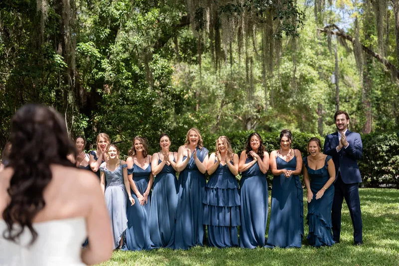 Giselle in white dress smiles while surrounded by bridesmaids in varying shades of blue under Spanish moss trees.