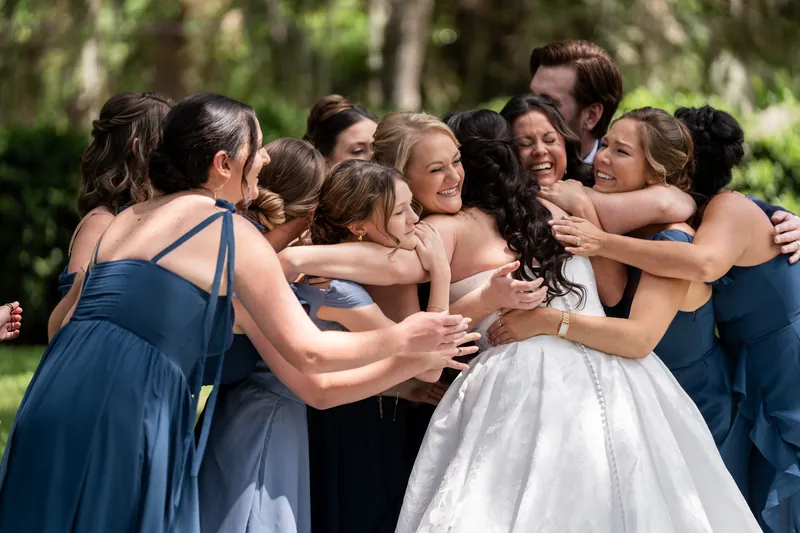 Bridesmaids in blue dresses huddle around Giselle in white dress for group embrace outdoors.