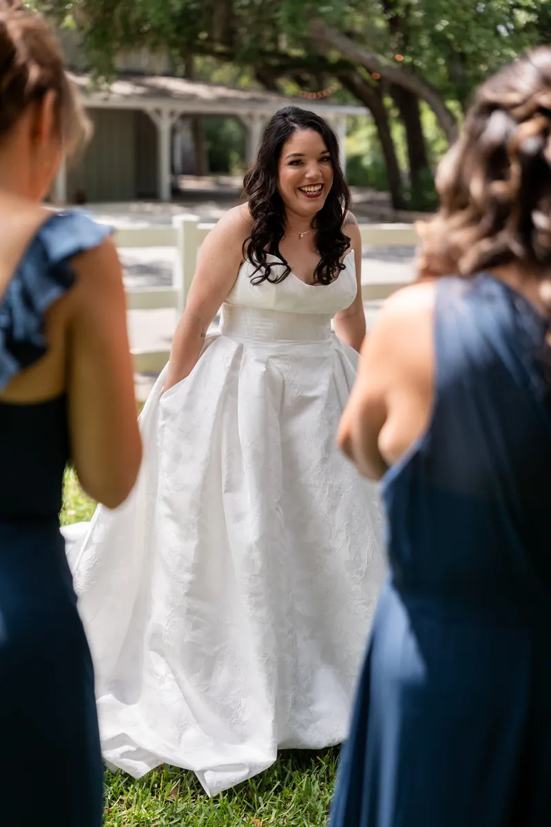 Giselle laughs with bridesmaids in blue dresses on grass, holding her white wedding dress skirt.