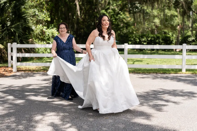 Giselle twirls her flowing white gown while bridesmaid holds the train on a tree-lined driveway.