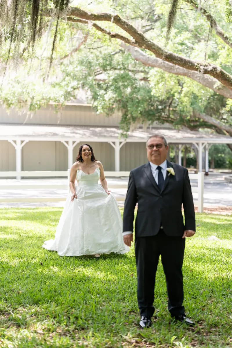 Duncan in black suit and Giselle in white strapless gown stand together on grass beneath sprawling oak tree.