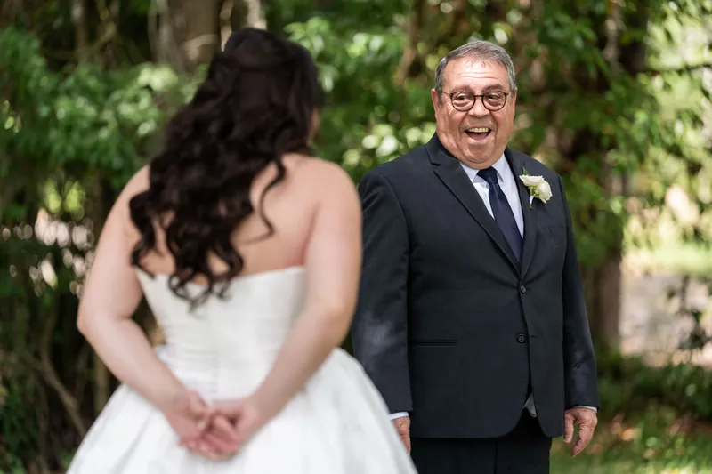 Giselle's father in dark suit with boutonnière smiles during first look with Giselle in white gown.