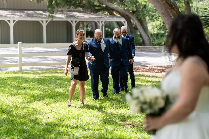 Giselle walks toward Duncan and groomsmen on a lawn near a white pavilion.