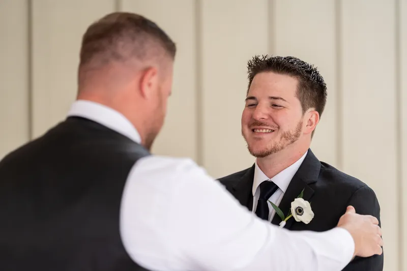 Duncan smiles while embracing a groomsman in formal black suits and white dress shirt indoors.