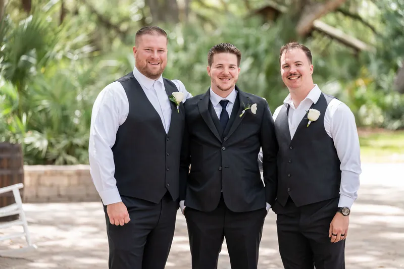 Duncan stands between two groomsmen in black and white formal wear with boutonnieres under trees.