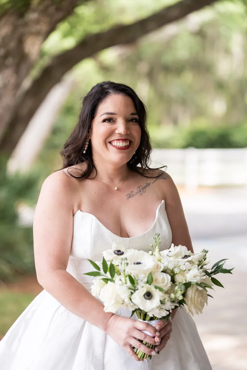 Giselle in white wedding gown holds white and green bouquet, smiling beneath large oak tree.