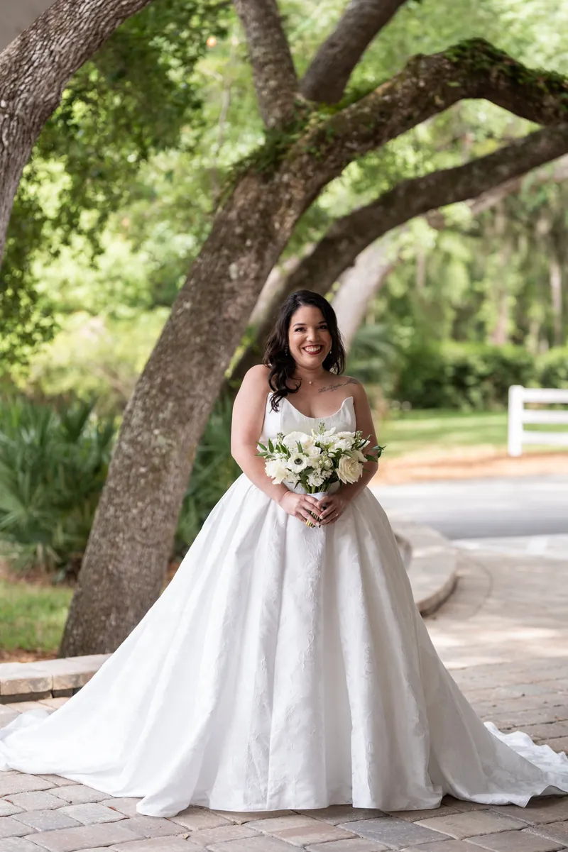 Giselle in ball gown wedding dress holds bouquet under sprawling oak tree on brick pathway.