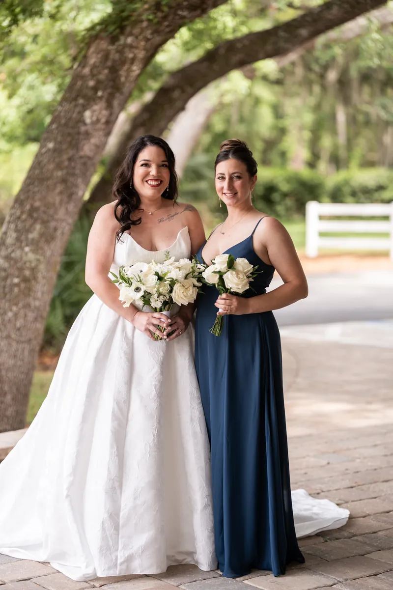 Giselle in white gown and bridesmaid in navy dress hold white bouquets under oak tree.