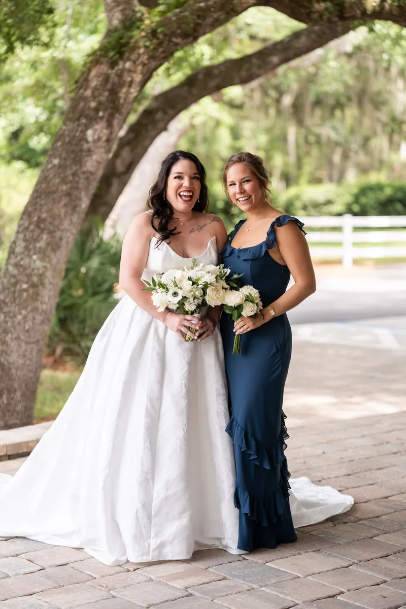 Giselle and bridesmaid pose together under oak tree, holding white bouquets on brick walkway.