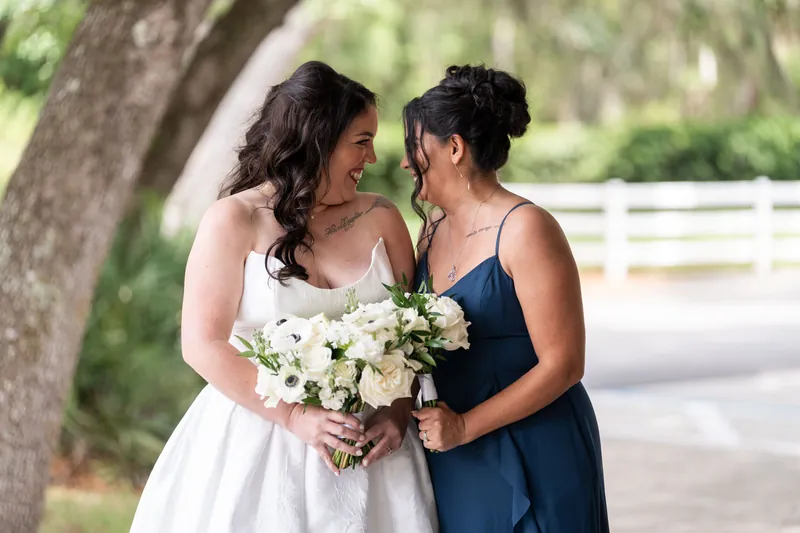 Giselle in white wedding dress and her companion in navy hold a white floral bouquet under a large tree.