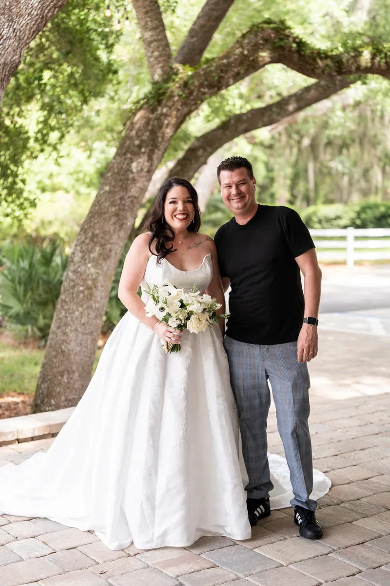 Duncan in black shirt and gray pants stands with Giselle in her white wedding gown holding white flowers by the tree.