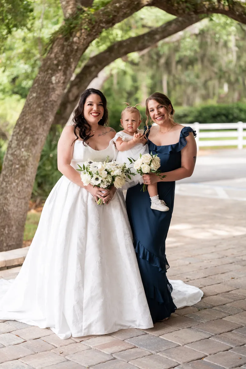 Giselle holds her white floral bouquet with two women in navy dresses, one holding a small child on the brick patio.