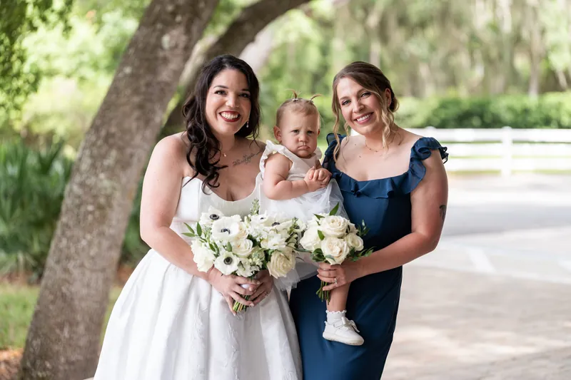 Giselle in white gown stands with two women in navy and a small child between them, all holding white flowers under the tree.