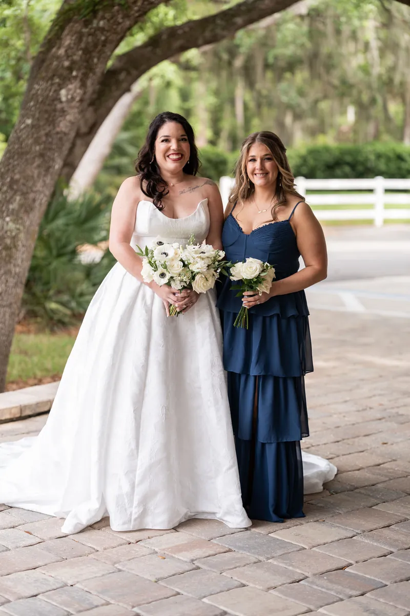 Giselle in white wedding dress and a woman in tiered navy dress hold matching white floral bouquets on the brick pathway.