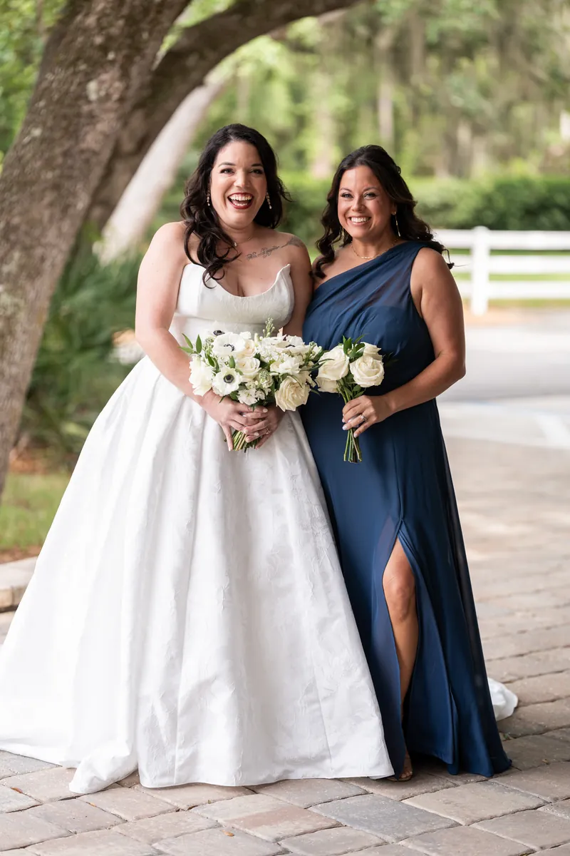 Giselle smiles at the camera while holding her bouquet next to a woman in slit navy dress holding white flowers under the tree.