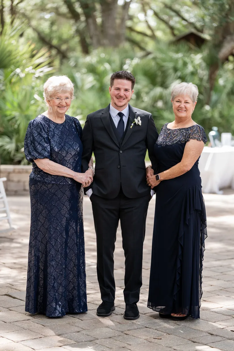 Duncan stands between two women in dark formal dresses under oak trees on a paved courtyard.
