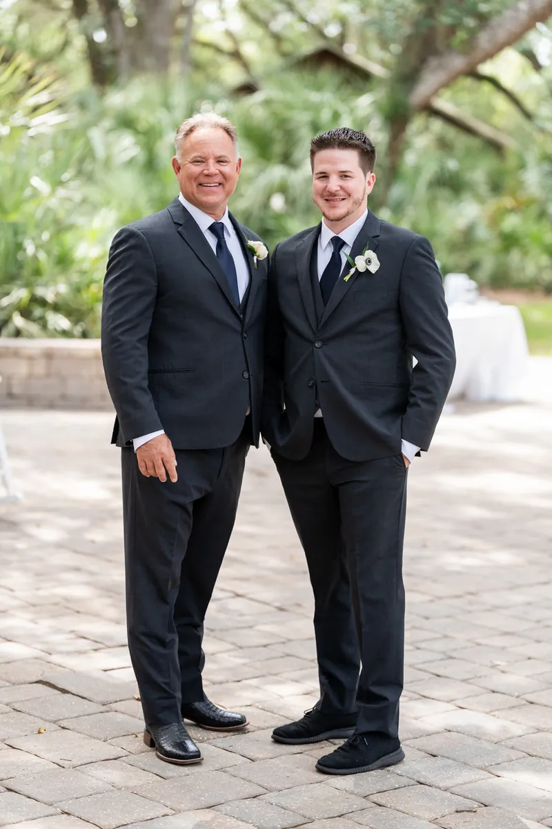 Duncan and his father stand together in dark suits with boutonnières under leafy trees.