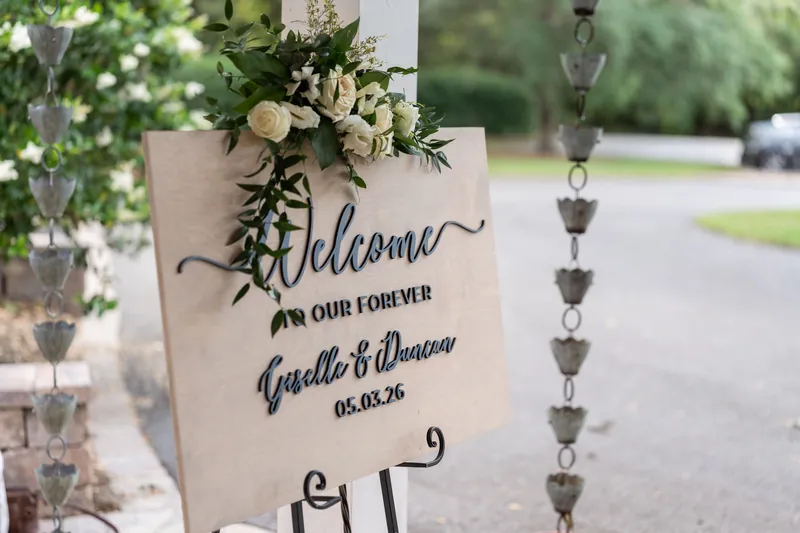 Cream welcome sign with white flowers reads "Welcome to our forever, Giselle & Duncan 05.03.26.