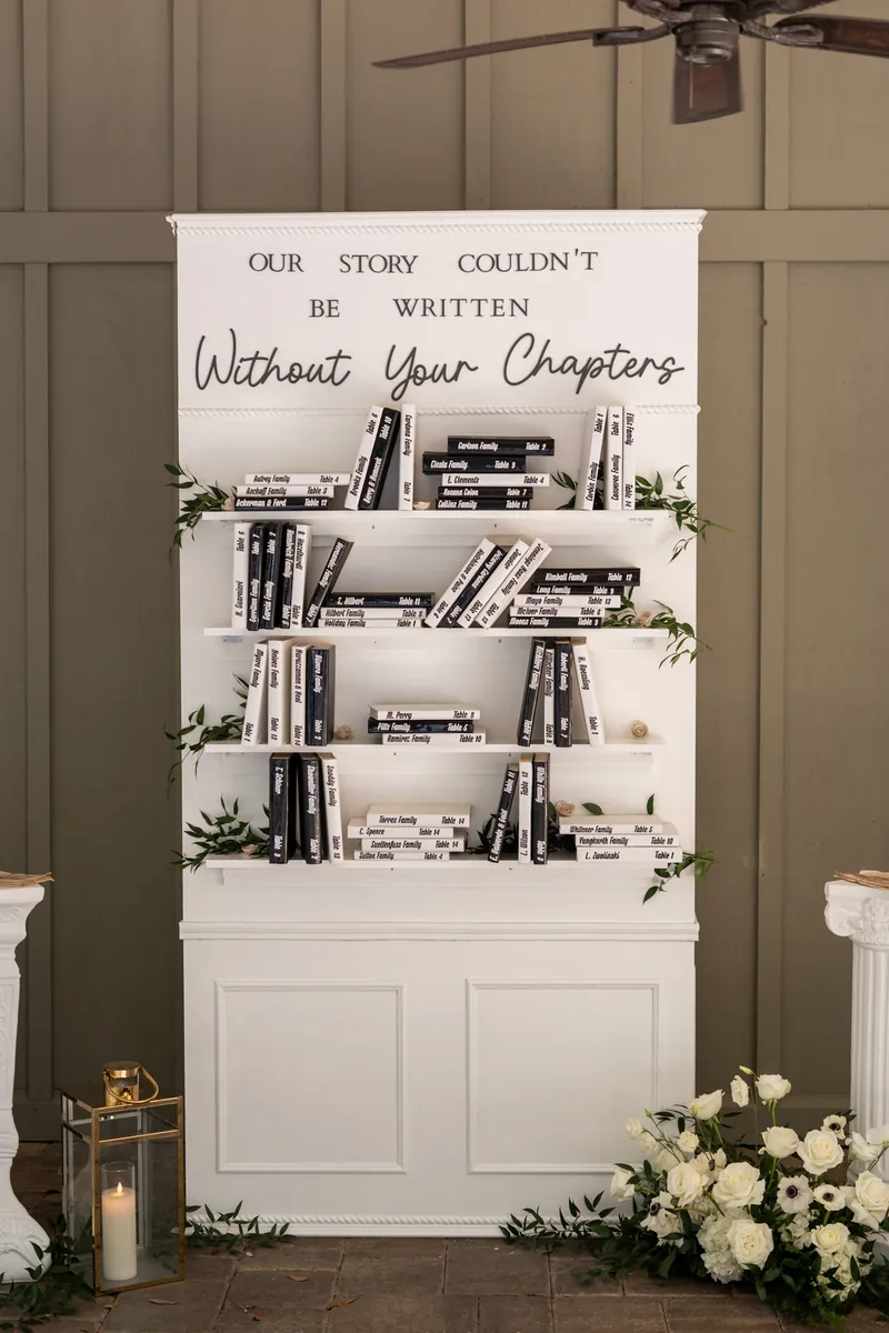 White bookshelf display reads "Our story couldn't be written without your chapters" with white flowers below.