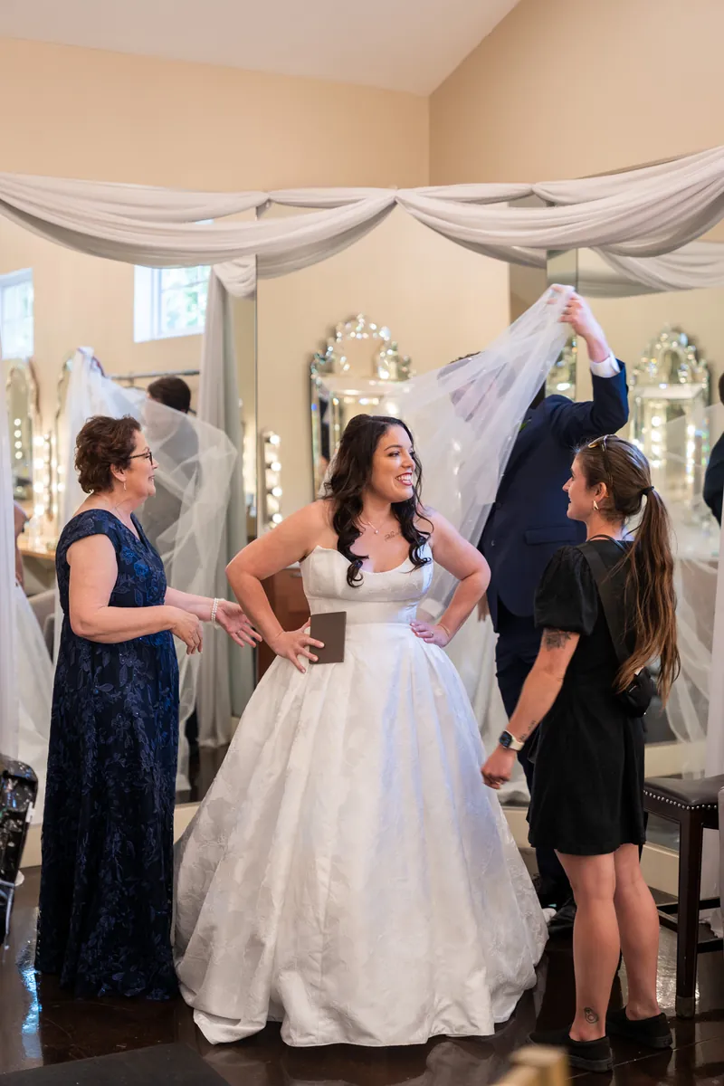 Giselle in a white wedding dress gets fitted by two women in a bridal shop with draped fabric overhead.