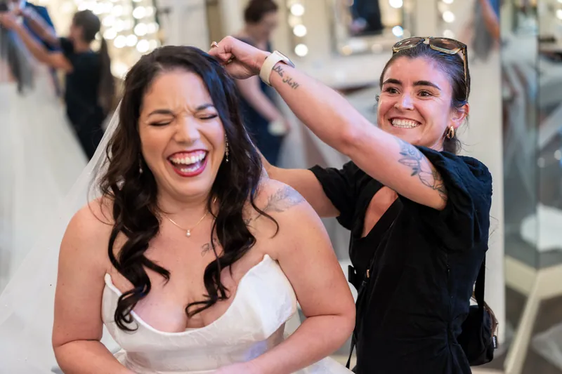 Giselle laughs while a bridesmaid adjusts her hair in front of a mirror at the bridal salon.