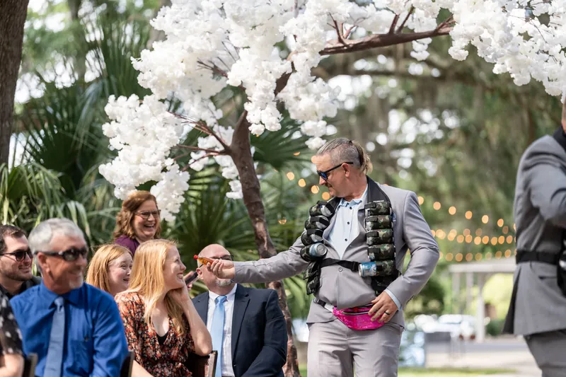 Duncan in a gray suit with pink bow tie speaks under white flowering trees at the outdoor wedding ceremony.