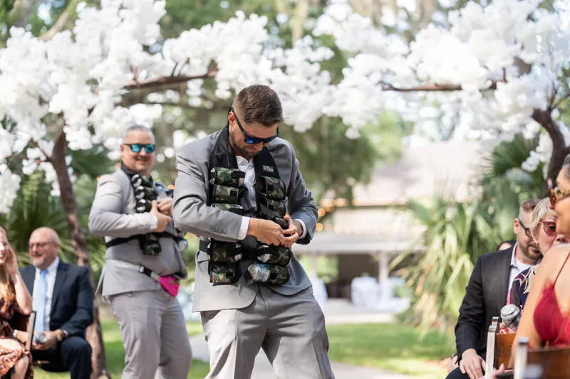 Duncan holds a camera under white blooming trees while wedding guests sit in folding chairs nearby.