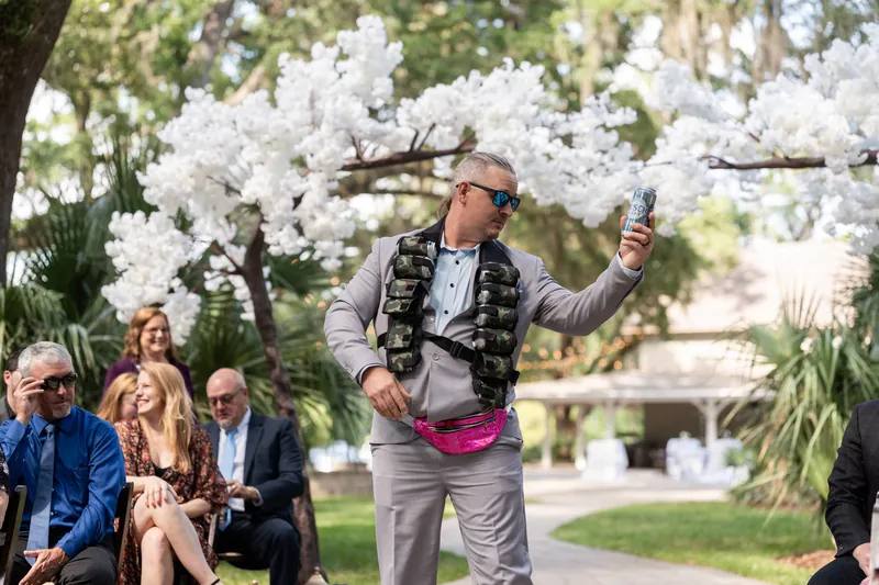 Duncan gestures while speaking under white flowering trees with seated guests and palm plants in background.