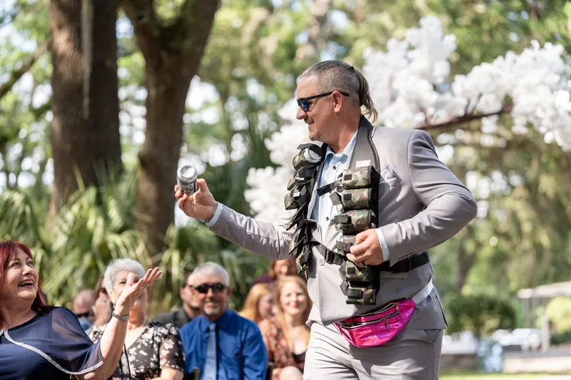 Duncan in gray suit and pink bow tie speaks animatedly with a microphone under white flowering trees.