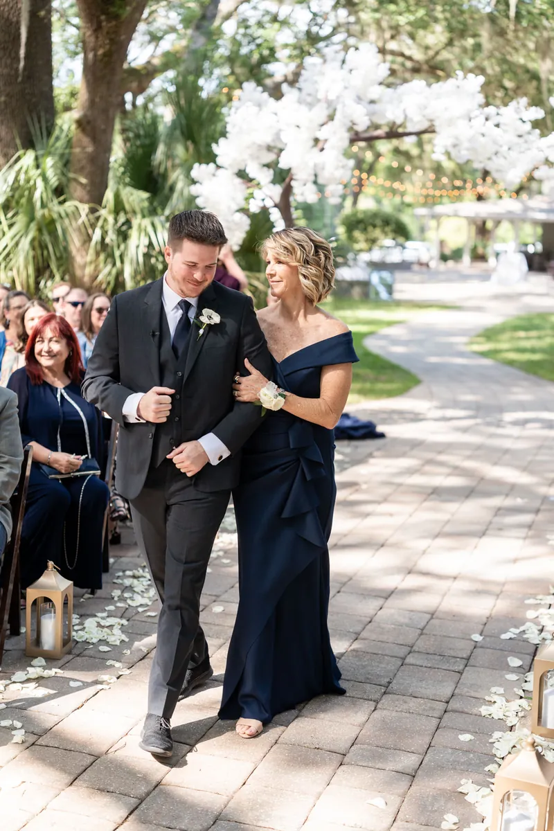 Duncan walks arm-in-arm with two women in black dresses down a tree-lined brick path.