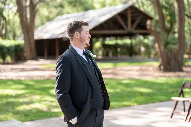 Duncan stands alone on a brick pathway wearing a dark suit, looking to the side near wooden structures.