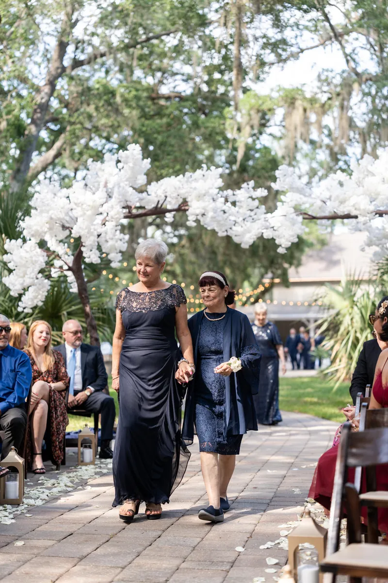Two women in dark dresses walk together beneath white flowering branches at an outdoor wedding venue.
