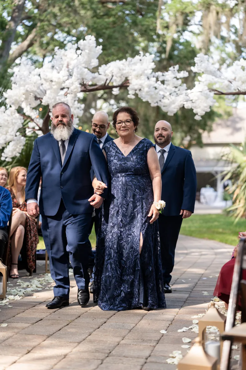 Four people walk down a brick aisle under white flowering branches, with guests seated on either side.
