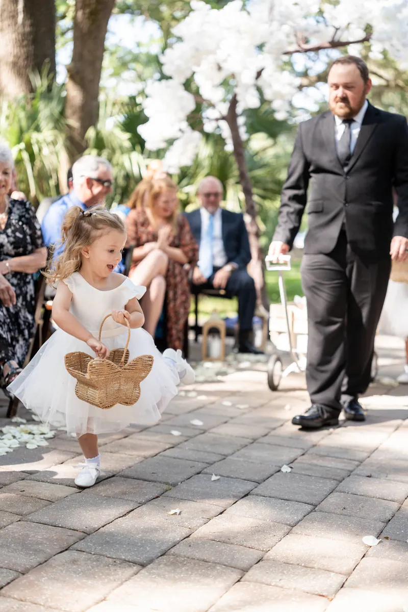 Young flower girl in white dress carries small basket while walking down aisle as Duncan watches from afar.