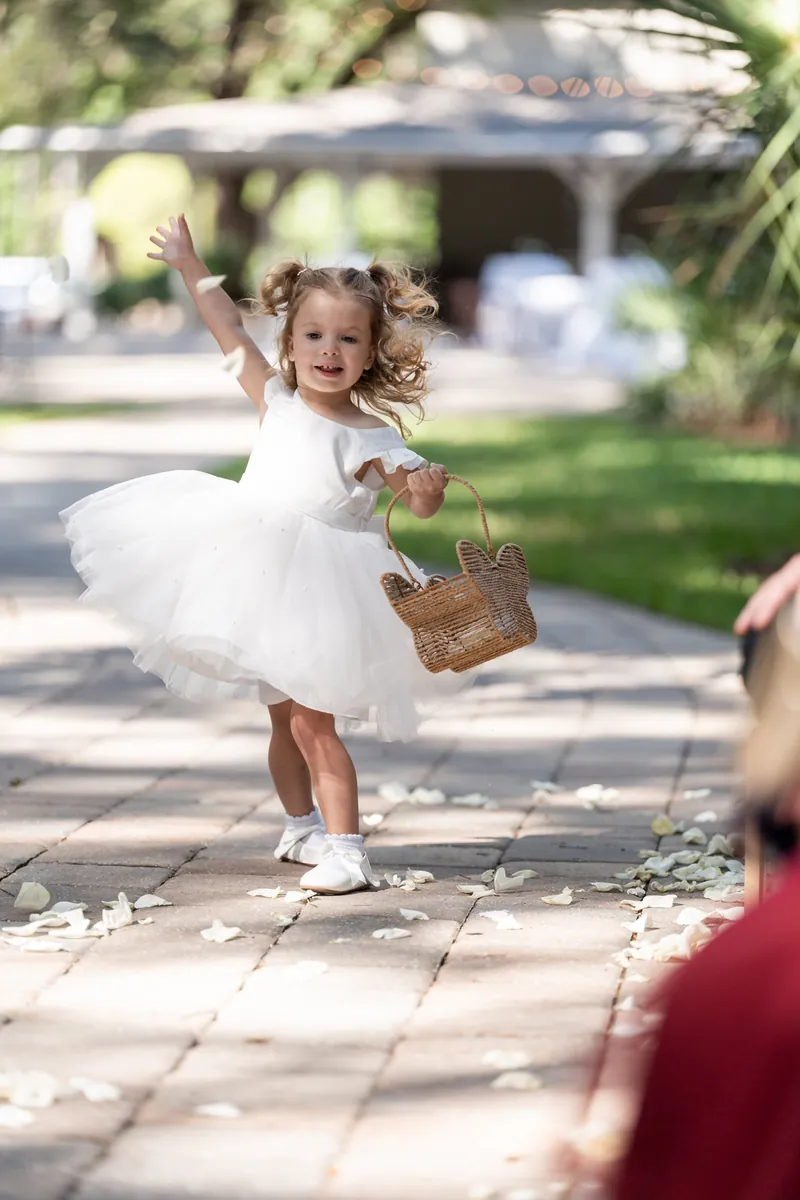 Flower girl in tulle skirt tosses petals from wicker basket during outdoor ceremony.
