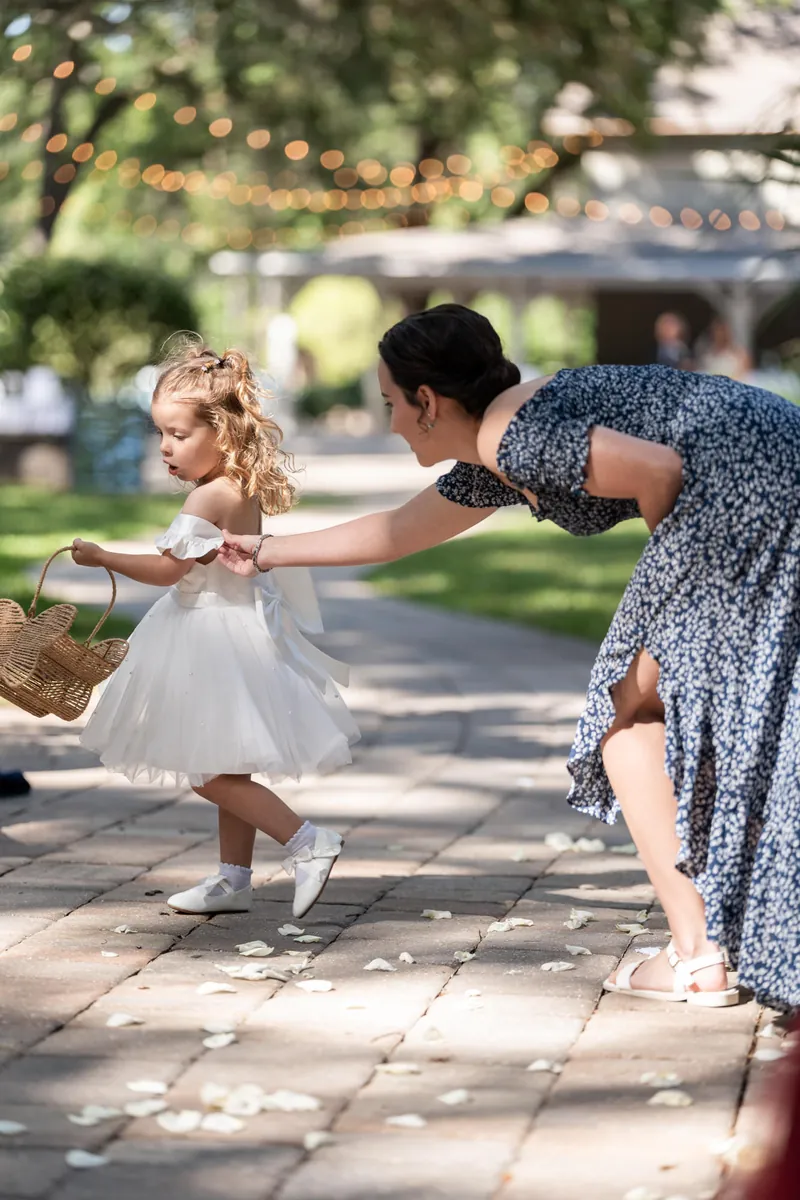 Woman helps toddler in white dress walk along stone pathway lined with wedding guests.